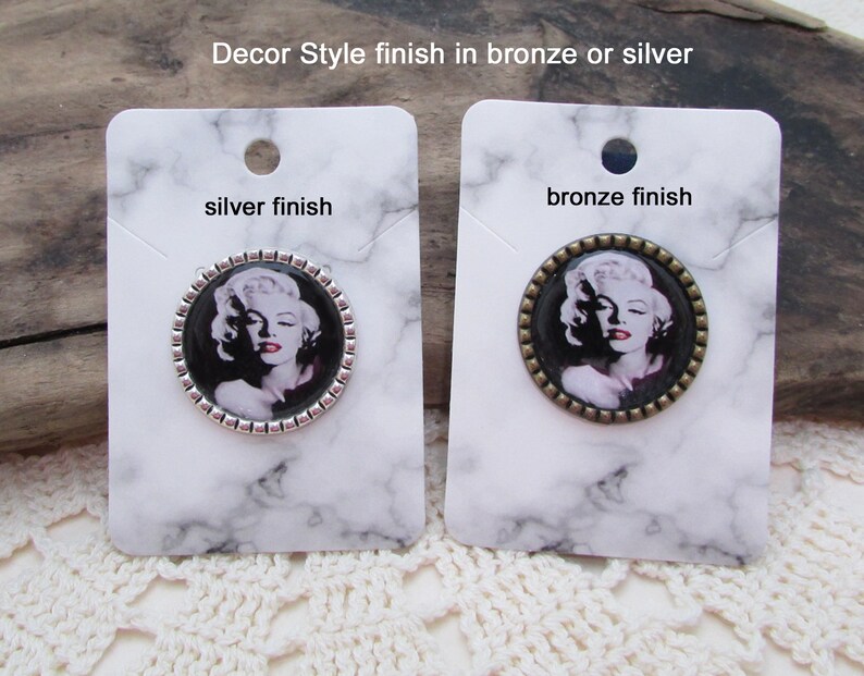 May include: Two round, silver and bronze, decorative brooches with a black and white image of Marilyn Monroe. The text "silver finish" and "bronze finish" is printed on the cards.