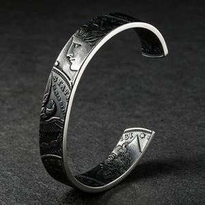 May include: A silver-toned cuff bracelet featuring a detailed relief design of historical coins. The surface displays embossed profiles of faces, an eagle, and the text "STATES" and "192". The metal has a dark, oxidized finish in the recessed areas.
