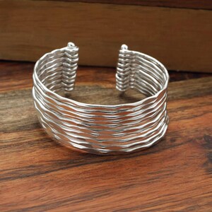 Puede incluir: Una pulsera de tipo brazalete ancha en color plata con un diseño de múltiples capas compuesto por varias bandas horizontales finas y onduladas, unidas en los dos extremos abiertos, creando un aspecto apilado con un acabado metálico pulido y brillante.