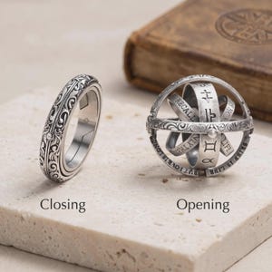 May include: A silver-toned metal ring featuring intricate scrollwork engravings. The ring is shown in two states: a closed band and an expanded spherical armillary structure with engraved text and zodiac symbols. The text reads Closing and Opening.