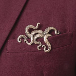 Pode incluir: Um broche de metal prateado em forma de polvo com múltiplos tentáculos curvos. A superfície da criatura apresenta um acabamento texturizado e martelado com pequenos detalhes circulares ao longo da parte inferior de cada braço, preso em tecido vermelho escuro.