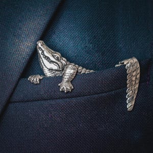 Può includere: Una spilla in metallo color argento a forma di coccodrillo con un dettagliato motivo a scaglie su testa, corpo e coda. Il coccodrillo è posizionato come se stesse uscendo da una tasca, con la testa e gli artigli anteriori visibili su un lato e la coda sull'altro.