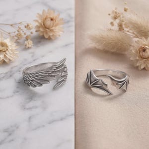 May include: Two silver-toned metal rings. One ring features a detailed design of feathered bird wings that wrap around the finger. The second ring features a design of smooth, pointed bat wings that also wrap around the finger in an open style.