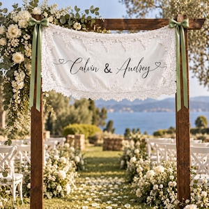 Può includere: Uno striscione in tessuto bianco con bordi in pizzo intricato, che presenta il testo stampato in nero "Calvin & Audrey" con piccole icone a forma di cuore. Lo striscione è appeso a una cornice di legno tramite due nastri di raso verde salvia.