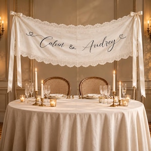 Può includere: Uno striscione in tessuto color crema con un intricato bordo in pizzo e lunghi nastri di raso a ciascuna estremità. Lo striscione mostra il testo in corsivo nero Calvin & Audrey, affiancato da piccole icone a forma di cuore, sopra un tavolo.
