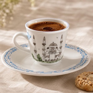 Peut inclure: Une tasse à expresso en céramique blanche avec sa soucoupe assortie, ornée d'une illustration peinte à la main représentant une mosquée avec des minarets dans des tons gris et verts. La soucoupe présente un motif de lignes bleues ondulées.