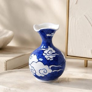 May include: A small, bulbous ceramic vase featuring a glossy finish with a deep blue background and white stylized cloud patterns. The vase has a narrow neck that flares out into a scalloped, wavy rim at the top, creating a distinct silhouette.