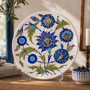 May include: A round ceramic decorative plate featuring a white background with a vibrant floral pattern in shades of deep blue, turquoise, and green. The design includes stylized blossoms and leafy stems arranged in a symmetrical composition.