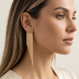 May include: A gold ear climber earring featuring a curved bar that sits along the earlobe, with multiple delicate gold chains of varying lengths hanging downward to create a cascading, asymmetrical fringe effect that rests against the neck.
