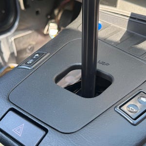 May include: A black automotive center console featuring a gear shift lever extending through a rectangular opening. The console includes a hazard light button, two window control switches, and a panel with the embossed text VAP near the shifter.