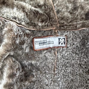 May include: A brown faux fur lined garment featuring a rectangular white fabric label with brown trim. The label displays the text manolo cremallera and a stylised logo of a person with arms raised. The interior lining has a textured appearance.