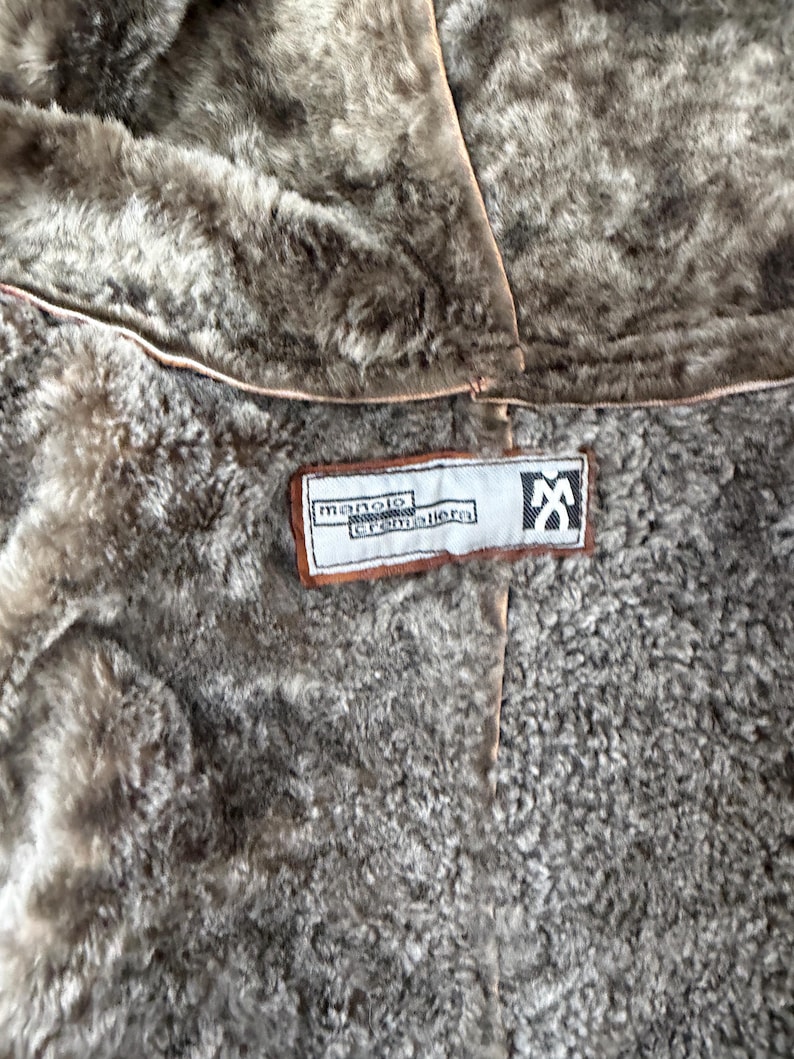 May include: A brown faux fur lined garment featuring a rectangular white fabric label with brown trim. The label displays the text manolo cremallera and a stylised logo of a person with arms raised. The interior lining has a textured appearance.