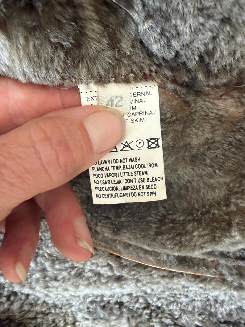 May include: A white garment care label with the number 42 printed in large font. The text includes instructions such as DO NOT WASH, COOL IRON, LITTLE STEAM, DO NOT USE BLEACH, and DO NOT SPIN. The label is attached to a textured brown faux fur lining.