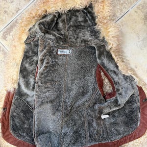 May include: A brown leather waistcoat with a thick, curly beige shearling trim along the edges and a dark grey faux fur lining. The interior features a rectangular label with text and a logo, and the garment is open to display the inner texture.