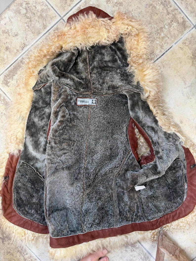 May include: A brown leather waistcoat with a thick, curly beige shearling trim along the edges and a dark grey faux fur lining. The interior features a rectangular label with text and a logo, and the garment is open to display the inner texture.