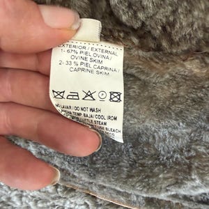 May include: A white garment care label with black text attached to a brown faux fur lining. The label lists 67% ovine skin and 33% caprine skin, along with laundry symbols and instructions including do not wash, cool iron, and do not use bleach.