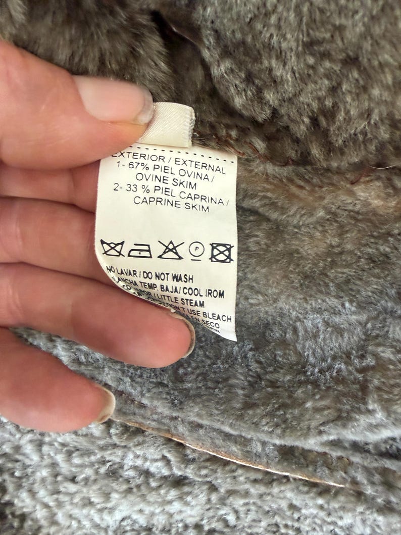 May include: A white garment care label with black text attached to a brown faux fur lining. The label lists 67% ovine skin and 33% caprine skin, along with laundry symbols and instructions including do not wash, cool iron, and do not use bleach.