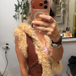 May include: A brown waistcoat featuring a thick, curly beige shearling trim along the front opening and armholes. The garment includes a small zipped pocket on the front with a circular metal pull tab, showcasing a textured, layered appearance.