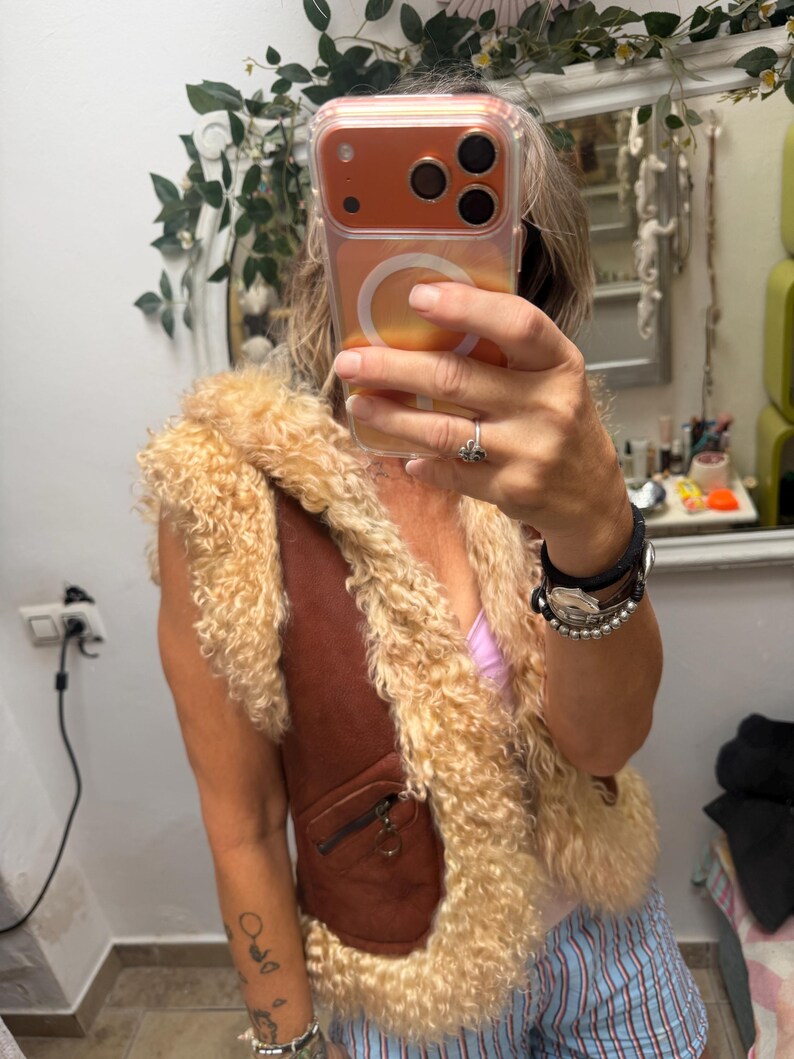 May include: A brown waistcoat featuring a thick, curly beige shearling trim along the front opening and armholes. The garment includes a small zipped pocket on the front with a circular metal pull tab, showcasing a textured, layered appearance.