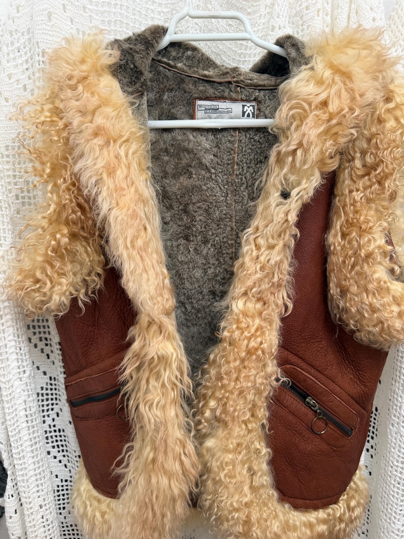 May include: A sleeveless waistcoat made of brown leather with a thick, curly, light beige shearling trim along the front edges, armholes, and hem. The interior is lined with dark grey fleece and features two front pockets with black zips.