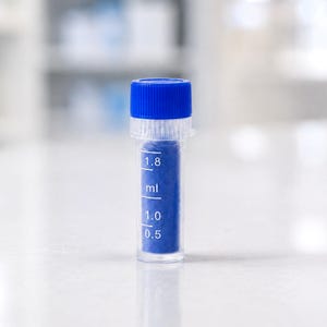 Puede incluir: Un pequeño vial de plástico transparente con una tapa de rosca azul brillante. El vial contiene una sustancia azul vibrante y presenta marcas de medición impresas en blanco que indican los niveles de 0,5, 1,0 y 1,8 ml en el recipiente.