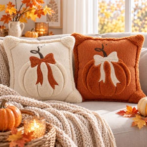 Peut inclure: Deux coussins carrés décoratifs présentant un motif de citrouille en relief et texturé. Le coussin de gauche est crème avec un nœud orange, et celui de droite est orange avec un nœud crème. Les deux ont une texture douce et duveteuse.