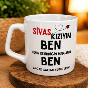 Pode incluir: Uma caneca de cerâmica branca com o texto em vermelho e preto SIVAS KIZIYIM BEN SENIN ESTIRDIGIN RÜZGARDA BEN ANCAK SACIMI KURUTURUM, apresentando um pequeno rastro de coração pontilhado e um gráfico de joaninha vermelha na frente.