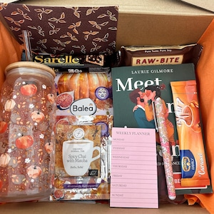Könnte beinhalten: Eine Geschenkbox mit einem Glasbecher mit Kürbismuster, dem Buch Meet Me in Maple Creek von Laurie Gilmore, einem rosa Wochenplaner, einem Blumenkugelschreiber, einer Packung Spicy Chai with Matcha Tee, Karamell-Getränkepulver und Riegeln.