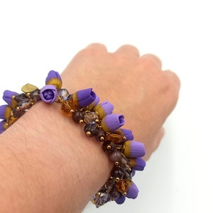 May include: A purple and gold beaded bracelet with small flower-shaped charms. The bracelet is on a person's wrist.