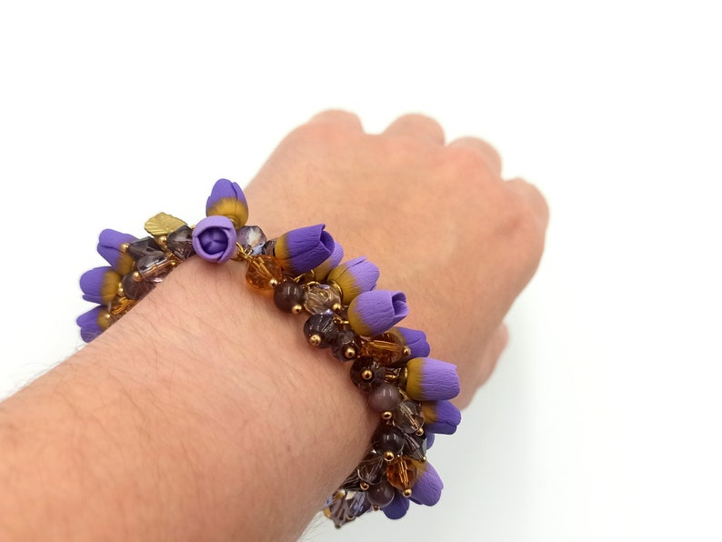 May include: A purple and gold beaded bracelet with small flower-shaped charms. The bracelet is on a person's wrist.