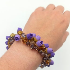 May include: A purple and gold beaded bracelet with flower-shaped charms. The bracelet is on a person's wrist.