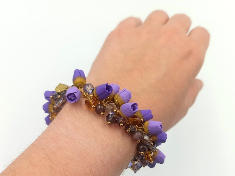 May include: A purple and gold beaded bracelet with flower-shaped charms. The bracelet is on a person's wrist.