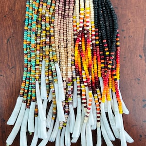 May include: A collection of beaded necklaces with various color schemes. The necklaces feature small, colorful beads in shades of turquoise, orange, black, and cream. Each necklace has a metal top and white, elongated pendants hanging from the bottom.