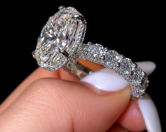 May include: A silver-toned ring featuring a large oval-cut center stone surrounded by a halo of small round stones. The band is encrusted with a continuous row of round stones along the visible side, all set in a metallic frame against a black background.