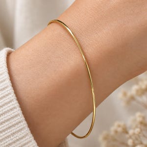 Può includere: Un sottile bracciale rigido in oro lucido con una superficie liscia e arrotondata. Il bracciale presenta un design circolare semplice e continuo che si adagia comodamente attorno al polso, mostrando una finitura metallica brillante e riflettente.