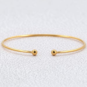 Op de afbeelding: Een goudkleurige open armband met een dunne, gepolijste metalen band die aan beide uiteinden eindigt in een kleine, gladde, bolvormige detail. Het ontwerp is eenvoudig en cirkelvormig, met een reflecterende, metalen afwerking.