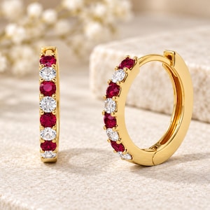 May include: A pair of gold hoop earrings featuring an alternating pattern of round-cut red rubies and clear white diamonds. The earrings have a hinged closure mechanism and are displayed upright on a textured surface.