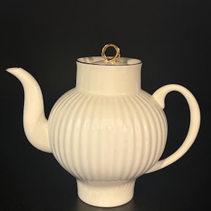 Op de afbeelding: Een witte keramische theepot met een verticaal geribbelde, ronde vorm, een gebogen tuit en een bijpassend deksel met een kleine, ronde goudkleurige knop. De theepot heeft een gladde, glanzende afwerking en een opvallende voet aan de onderzijde.
