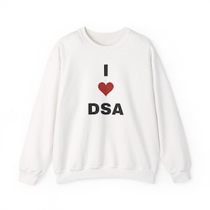 May include: A white long-sleeve crewneck sweatshirt featuring a graphic print on the chest. The design consists of the letter I in black, a red heart symbol, and the letters DSA in black, arranged vertically in the center of the garment.