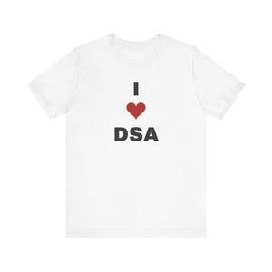 Può includere: Una t-shirt bianca a maniche corte con il testo I seguito da un simbolo di cuore rosso e le lettere DSA in maiuscolo nero centrate sul petto. Il capo presenta uno scollo rotondo e una vestibilità standard con maniche corte.