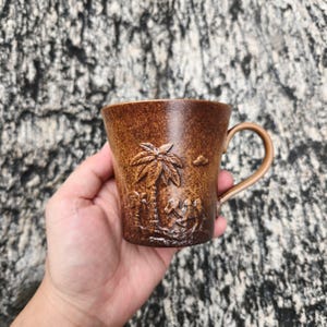 May include: A brown ceramic mug with a textured, speckled finish. The exterior features a raised, embossed design depicting a palm tree and a camel in a desert landscape. The mug has a curved handle on the right side and a tapered, conical shape.