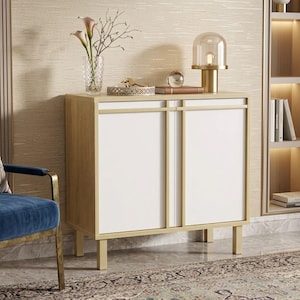 May include: A rectangular cabinet with a light wood grain frame and two white doors featuring gold-toned vertical trim. The top surface holds a clear glass vase with flowers, a decorative bowl, a small glass sphere, and a gold table lamp.