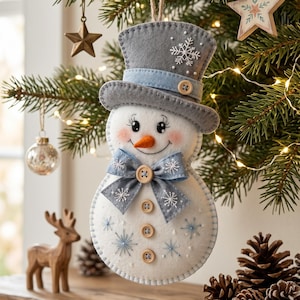 May include: A felt snowman ornament featuring a grey top hat with a light blue band, a carrot-shaped nose, and a light blue bow tie. The snowman is decorated with small wooden buttons, embroidered white snowflakes, and pink cheeks, hanging from a jute twine loop.
