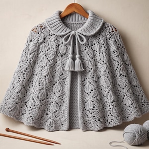 May include: A light gray knitted cape featuring an intricate leaf-like lace pattern, a ribbed collar, and a front tie closure with two matching tassels. The garment is displayed on a wooden hanger, accompanied by two balls of gray yarn and two knitting needles.