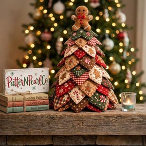 Pode incluir: Um enfeite de árvore de Natal de tecido construído com camadas de retalhos acolchoados em vermelho, verde e creme com padrões de bonecos de gengibre, bengalas doces e flocos de neve, rematado com uma pequena figura de boneco de gengibre.