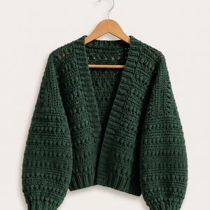 May include: A dark forest green crochet cardigan featuring a textured bobble stitch pattern, long sleeves with ribbed cuffs, and a wide ribbed front placket. The garment is displayed on a wooden hanger, with the text PatternNPearlCo in the corner.