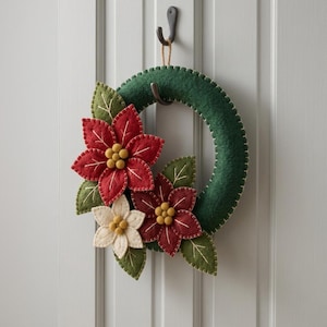 Op de afbeelding: Een ronde krans van donkergroen vilt, voorzien van twee grote rode vilten kerststerren en één kleinere witte vilten kerstster. Elke bloem heeft een hartje gemaakt van kleine gele vilten balletjes. De krans hangt aan een bruin touwtje.