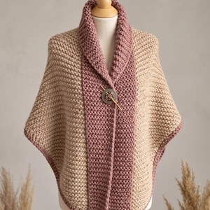 May include: A knitted poncho featuring a textured stitch pattern in beige and dusty rose. The garment has a wide shawl collar and is fastened at the center front with a circular, antique-style metal pin featuring an intricate embossed design.
