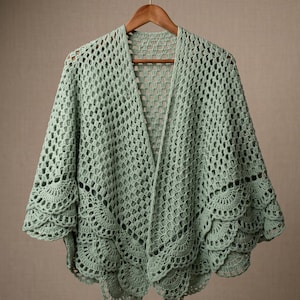 May include: A light sage green crochet shawl featuring an openwork grid pattern throughout the main body and a decorative scalloped edge along the bottom hem. The garment is displayed on a wooden hanger, showcasing its intricate yarn construction.