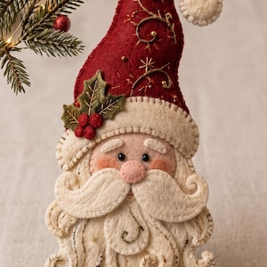 May include: A felt Santa Claus ornament featuring a deep red curved hat with gold embroidery and a white pom-pom. The figure has a cream-colored beard, mustache, and hat trim, accented with green holly leaves and red berries on the side.